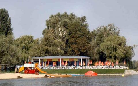 Ein Strand am See mit einem Spielplatz, einem Gebäude mit Sitzgelegenheiten im Freien und Reihen von orangefarbenen Sonnenschirmen, umgeben von Bäumen.