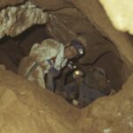 Drei Personen mit Helmen und Scheinwerfern erkunden einen engen, felsigen Höhlengang.