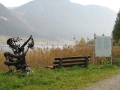 Eine Holzbank, eine Treibholzskulptur und ein Informationsschild stehen an einem See, im Hintergrund sind die Berge zu sehen.