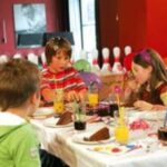 Kinder sitzen um einen Tisch herum, essen Kuchen und trinken Saft auf einer Geburtstagsfeier, im Hintergrund sind Partydekorationen und Bowlingkegel zu sehen.