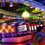 Die Leute sitzen an Tischen und einer Bar in einem lebhaften, farbenfrohen Nachtclub mit Neonlicht, einer Discokugel und modernem Dekor.