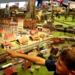 Zwei Kinder betrachten eine detailgetreue Modelleisenbahnanlage mit Miniaturgebäuden, -fahrzeugen und -gleisen, die im Haus ausgestellt ist, und zeigen auf sie.