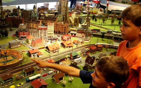 Zwei Kinder betrachten eine detailgetreue Modelleisenbahnanlage mit Miniaturgebäuden, -fahrzeugen und -gleisen, die im Haus ausgestellt ist, und zeigen auf sie.