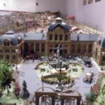Modellbahnhof mit detaillierten Gebäuden, einem Springbrunnen, Pferdekutschen, Bäumen und Miniaturfiguren in einem naturgetreuen Diorama.