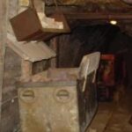 In einem schwach beleuchteten Tunnel mit steinernen Wänden und einer schrägen Decke befinden sich ein Förderwagen und eine Ausrüstung.