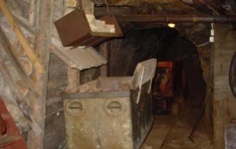 In einem schwach beleuchteten Tunnel mit steinernen Wänden und einer schrägen Decke befinden sich ein Förderwagen und eine Ausrüstung.