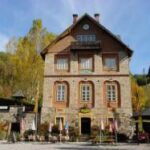 Ein dreistöckiges Gebäude aus Stein und Holz mit symmetrischen Fenstern, einem Spitzdach und Fahnen am Eingang vor einer Kulisse aus Bäumen und blauem Himmel.