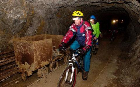 Menschen mit Helmen und Jacken fahren auf Fahrrädern in einem schwach beleuchteten Tunnel neben einem alten Bergbauwagen auf Schienen.
