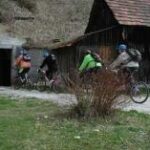 Vier Personen mit Helmen und Rucksäcken fahren auf Fahrrädern einen Weg entlang, der an einem rustikalen Holzgebäude und einem Busch vorbeiführt.