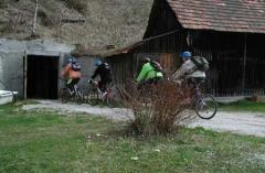 Vier Personen mit Helmen und Rucksäcken fahren auf Fahrrädern einen Weg entlang, der an einem rustikalen Holzgebäude und einem Busch vorbeiführt.