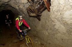 Zwei Radfahrer, die Helme und Sicherheitsausrüstung tragen, fahren mit ihren Fahrrädern durch einen schwach beleuchteten, felsigen Tunnel, über dem sich Bergbaumaschinen befinden.