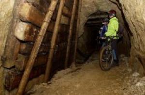 Zwei Personen mit Helmen und Sicherheitsausrüstung stehen mit ihren Fahrrädern in einem schwach beleuchteten, verstärkten Tunnel mit Holzstützen.