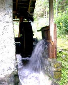 Ein hölzernes Wasserrad, das an einem steinernen Gebäude befestigt ist, wird durch fließendes Wasser in einem bewaldeten Außenbereich gedreht.