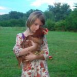 Ein junges Mädchen steht auf einer Wiese und hält ein braunes Huhn in den Armen. Im Hintergrund sind Bäume und ein Spielzeuglastwagen zu sehen.