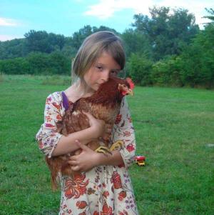 Ein junges Mädchen steht auf einer Wiese und hält ein braunes Huhn in den Armen. Im Hintergrund sind Bäume und ein Spielzeuglastwagen zu sehen.