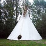 Ein großes weißes Tipi steht auf einer Wiese, umgeben von Bäumen, mit einer Öffnung an der Vorderseite und Holzstangen an der Spitze.