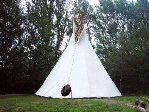 Ein großes weißes Tipi steht auf einer Wiese, umgeben von Bäumen, mit einer Öffnung an der Vorderseite und Holzstangen an der Spitze.
