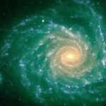 Eine helle Spiralgalaxie mit leuchtenden Armen strahlt von einem leuchtenden Zentrum vor einem dunklen, grünlichen Hintergrund aus, wobei links unten ein kleineres Objekt sichtbar ist.