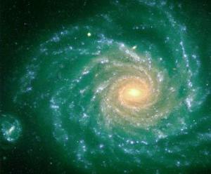 Eine helle Spiralgalaxie mit leuchtenden Armen strahlt von einem leuchtenden Zentrum vor einem dunklen, grünlichen Hintergrund aus, wobei links unten ein kleineres Objekt sichtbar ist.