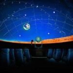 Sitzreihen mit Blick auf eine Kuppel mit einer projizierten Sternenkarte und Sternbildern in einem dunklen Planetarium.