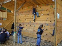 Kinder mit Klettergurten klettern an einer Indoor-Kletterwand aus Holz, während andere in der Nähe warten. Der Bereich ist überdacht und mit einem Holzspanboden versehen.