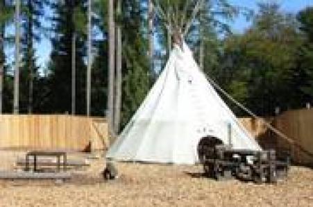 Ein weißes Tipi aus Segeltuch steht auf Holzspänen neben Picknicktischen und einem Holzzaun, umgeben von Bäumen.