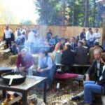 Eine Gruppe von Menschen sitzt und steht um Picknicktische im Freien, während im Vordergrund eine große Pfanne über einem offenen Feuer kocht. Im Hintergrund sind Bäume und ein Holzzaun zu sehen.