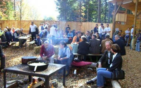 Eine Gruppe von Menschen sitzt und steht um Picknicktische im Freien, während im Vordergrund eine große Pfanne über einem offenen Feuer kocht. Im Hintergrund sind Bäume und ein Holzzaun zu sehen.