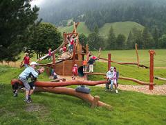 Kinder spielen auf einem hölzernen Spielplatz in einem grasbewachsenen Außenbereich mit Bäumen und Hügeln im Hintergrund.