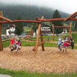 Kinder spielen auf Holzschaukeln auf einem Spielplatz im Freien, umgeben von Gras, Bäumen und Häusern, mit Bergen und Nebel im Hintergrund.