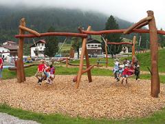Kinder spielen auf Holzschaukeln auf einem Spielplatz im Freien, umgeben von Gras, Bäumen und Häusern, mit Bergen und Nebel im Hintergrund.