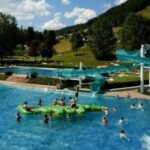 Freibad mit Kindern, die auf einer grünen aufblasbaren Struktur spielen und eine blaue Wasserrutsche benutzen; grasbewachsene Hügel und Bäume im Hintergrund.