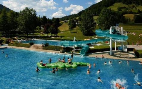 Freibad mit Kindern, die auf einer grünen aufblasbaren Struktur spielen und eine blaue Wasserrutsche benutzen; grasbewachsene Hügel und Bäume im Hintergrund.
