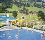 Menschen schwimmen in einem Freibad mit einer blauen Rutsche, umgeben von Grashügeln, Bäumen und verstreuten Sonnenanbetern. Im Hintergrund sind Gebäude zu sehen.