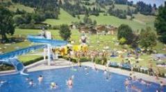 Menschen schwimmen in einem Freibad mit einer blauen Rutsche, umgeben von Grashügeln, Bäumen und verstreuten Sonnenanbetern. Im Hintergrund sind Gebäude zu sehen.