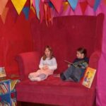 Zwei Kinder sitzen auf einem übergroßen roten Stuhl in einem farbenfrohen, mit Flaggen geschmückten Raum; ein Kind liest ein Buch, während das andere daneben sitzt. In der Nähe sind Bücher in Regalen ausgestellt.
