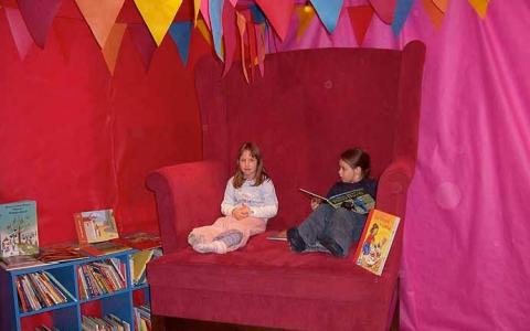 Zwei Kinder sitzen auf einem übergroßen roten Stuhl in einem farbenfrohen, mit Flaggen geschmückten Raum; ein Kind liest ein Buch, während das andere daneben sitzt. In der Nähe sind Bücher in Regalen ausgestellt.