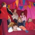 Sechs Kinder sitzen und lümmeln auf einem großen roten Stuhl in einem Zelt, das mit bunten dreieckigen Wimpeln geschmückt ist.