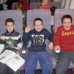 Drei Jungen sitzen zusammen in flugzeugähnlichen Sitzen; einer hält ein Buch, einer winkt und lächelt, ein anderer grinst breit. Hinter ihnen sitzen andere Kinder, die den Blick abwenden.