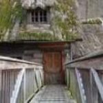 Ein hölzerner Steg führt zur Eingangstür eines alten Hauses, an dessen Dach und Wänden Moos und Pflanzen wachsen.