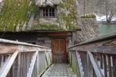 Ein hölzerner Steg führt zur Eingangstür eines alten Hauses, an dessen Dach und Wänden Moos und Pflanzen wachsen.