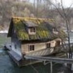 Ein kleines Haus mit moosbewachsenem Dach steht am Rande eines Flusses, der über einen Holzsteg mit dem Ufer verbunden ist.