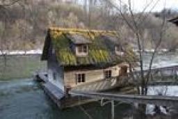 Ein kleines Haus mit moosbewachsenem Dach steht am Rande eines Flusses, der über einen Holzsteg mit dem Ufer verbunden ist.