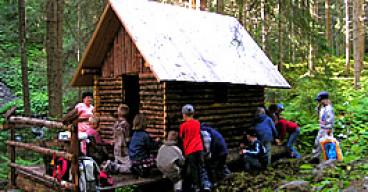 Eine Gruppe von Kindern und ein Erwachsener sitzen und stehen in der Nähe einer kleinen Holzhütte in einem Waldgebiet.
