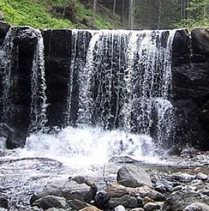 Ein kleiner Wasserfall fließt über eine Steinmauer in einen felsigen Bach, umgeben von Grün und Bäumen im Hintergrund.