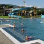 Kinder schwimmen in einem Freibad mit einer Wasserrutsche und einem pilzförmigen Springbrunnen. Bei klarem Himmel sind im Hintergrund Gebäude und Hügel zu sehen.