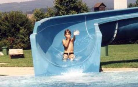 Ein Kind rutscht eine blaue Wasserrutsche hinunter und spritzt am Ende in ein Becken. Im Hintergrund sind Gras und Bäume zu sehen.