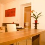 Ein moderner Essbereich mit einem Holztisch, weißen Stühlen, einem roten Gemälde an der Wand und einer Topfpflanze auf einem kleinen Sockel.