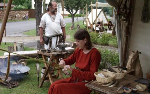 Eine Frau in historischer Kleidung arbeitet mit Werkzeugen an einem Holztisch im Freien, während ein Mann in der Nähe steht; andere Personen sind im Hintergrund zu sehen.