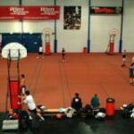 In einer Sporthalle wird auf mehreren Plätzen Basketball gespielt, wobei Bänke und Sportgeräte den Rand des Spielfelds säumen.